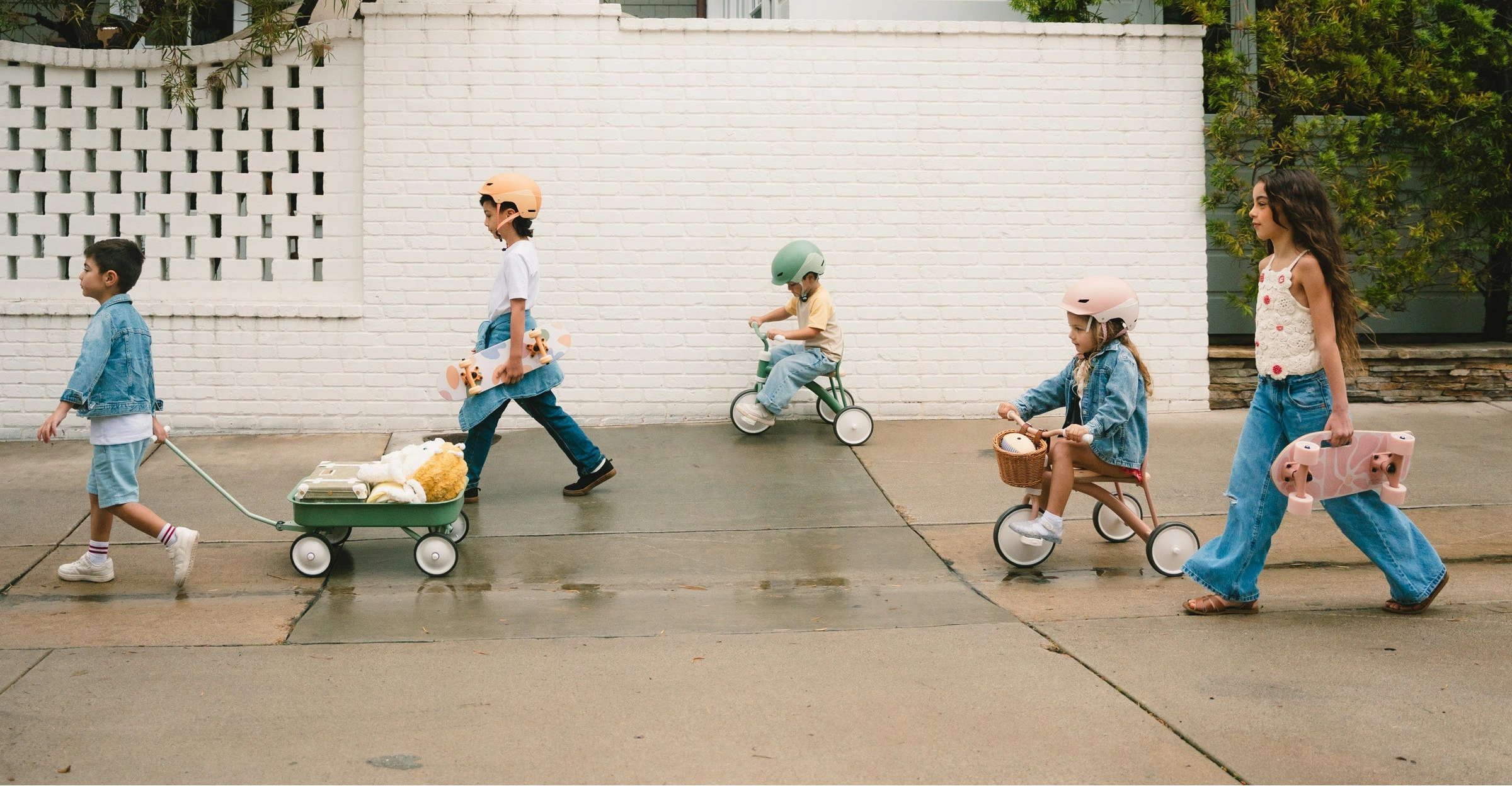 Five children playing with various toys on a sidewalk in front of a white building.