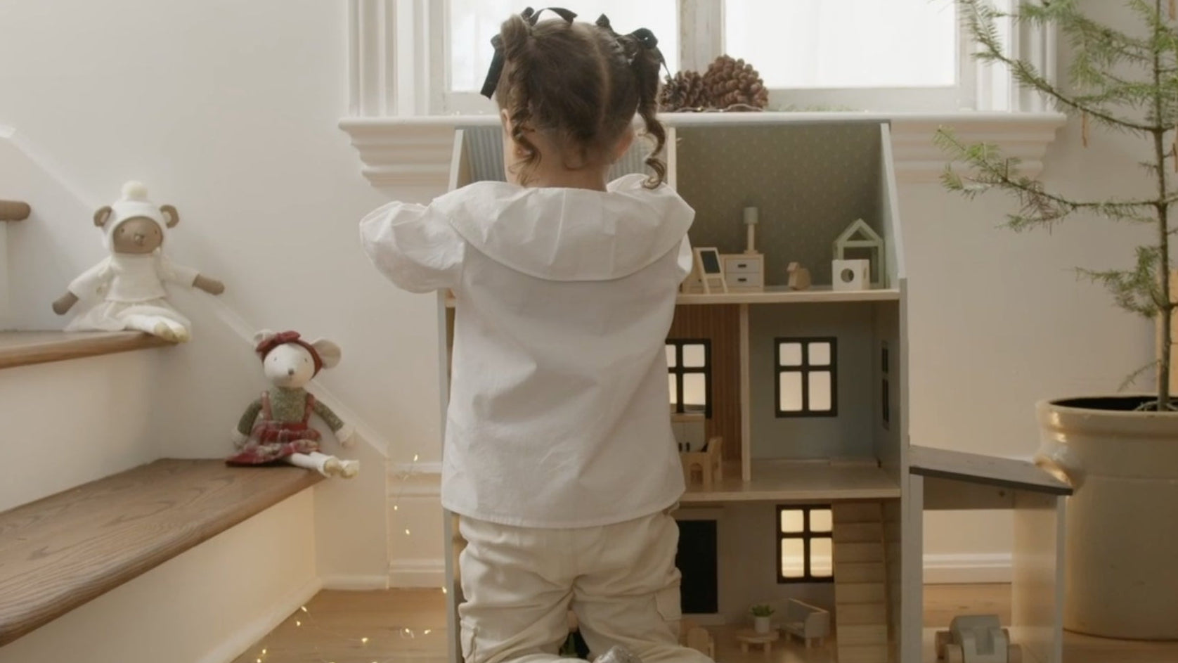 Child playing with a dollhouse in a bright room with toys and plants.